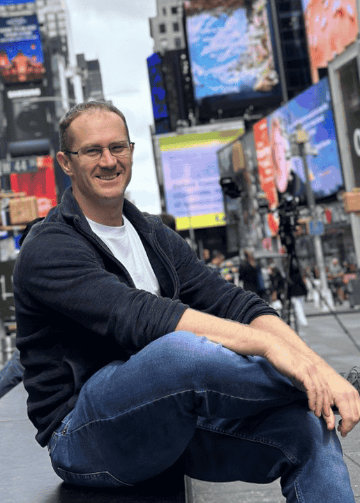 Fantasy author James Brennan sits casually on the steps in a bustling city, surrounded by bright billboards that could easily serve as inspiration for his next magical saga.